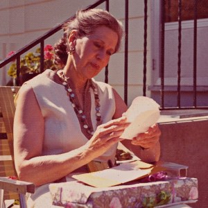 An older Genevieve, seated near a house, reads her birthday card at a party for her in Minneapolis